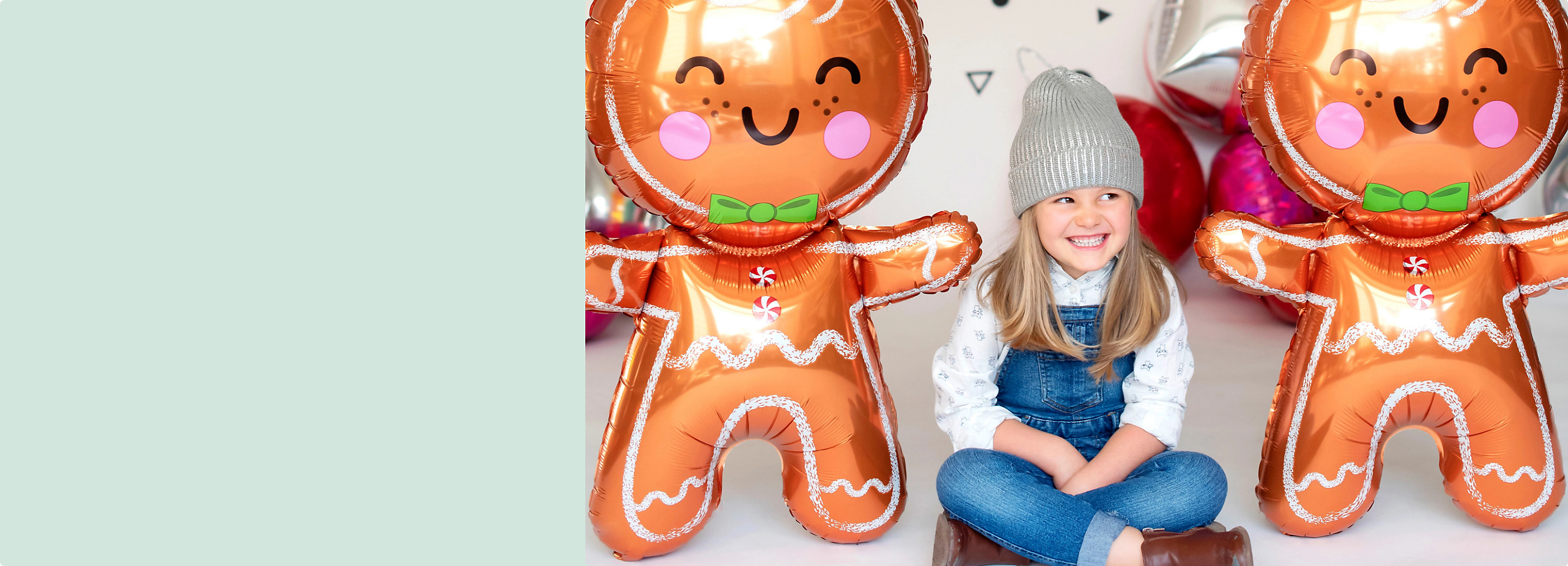 A smiling girl sitting on a floor between two freestanding giant gingerbread man foil balloons. 