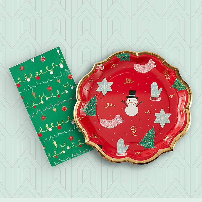 A red Sophistiplate Deck the Halls plate with Christmas-themed print and a Sophistiplate Deck the Halls guest towel with a green print. 