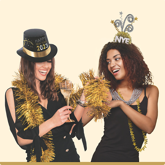 Two women wearing NYE accessories including a “Happy New Year 2023” top hat, a happy new year bead necklace and a gold tinsel boa.