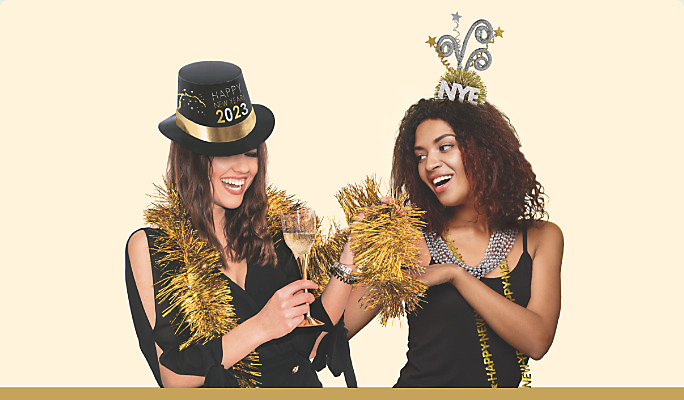 Two women wearing NYE accessories including a “Happy New Year 2023” top hat, a happy new year bead necklace and a gold tinsel boa.
