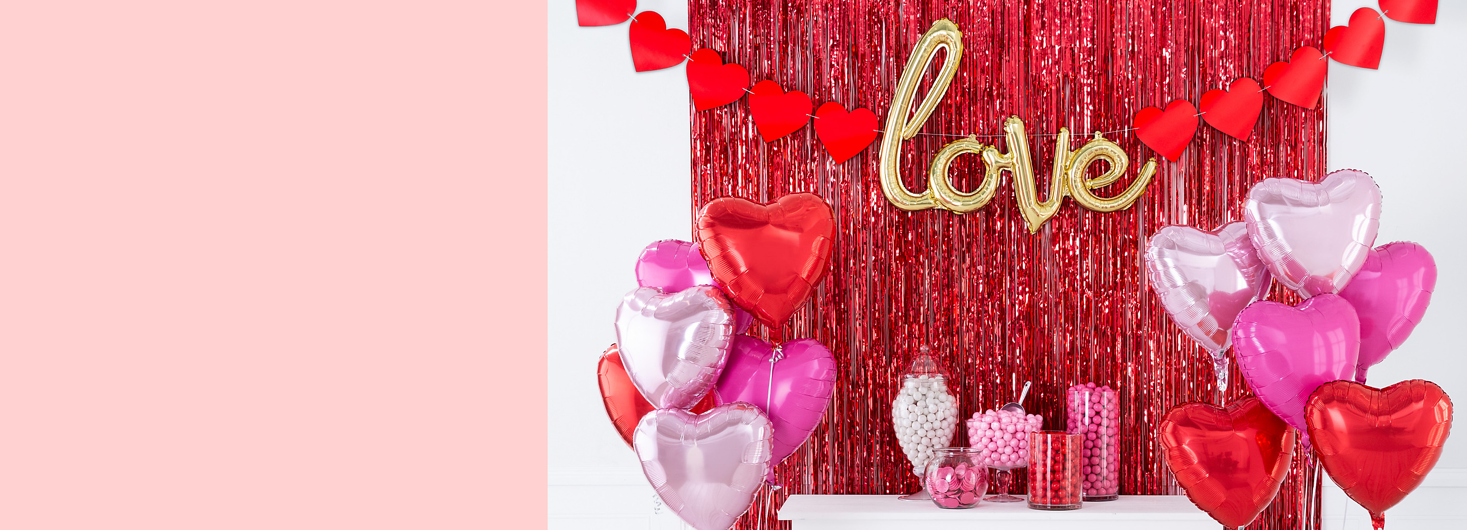 A room styled with a red tinsel curtain, a gold love balloon banner, a candy buffet and two pink and red heart foil balloon bouquets. 