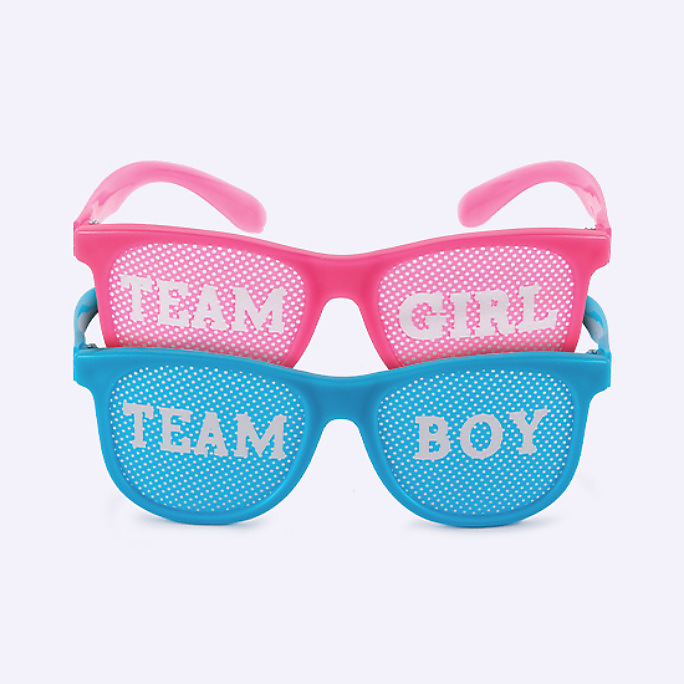 Pink plastic party glasses with lenses printed with “team girl” stacked with a pair of blue glasses with “team boy” lenses.