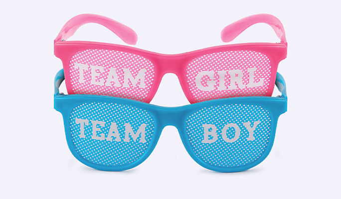 Pink plastic party glasses with lenses printed with “team girl” stacked with a pair of blue glasses with “team boy” lenses.