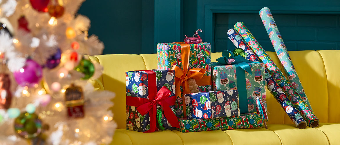 Une collection de cadeaux de Noël emballés avec éclat et des rouleaux de papier d’emballage assortis sur un canapé jaune à côté d’un arbre de Noël blanc décoré. Du papier d’emballage coloré, des motifs festifs, des rubans et des nœuds créent une présentation de cadeaux festifs et gais, mettant en vedette le papier et l’emballage coordonnés. 
