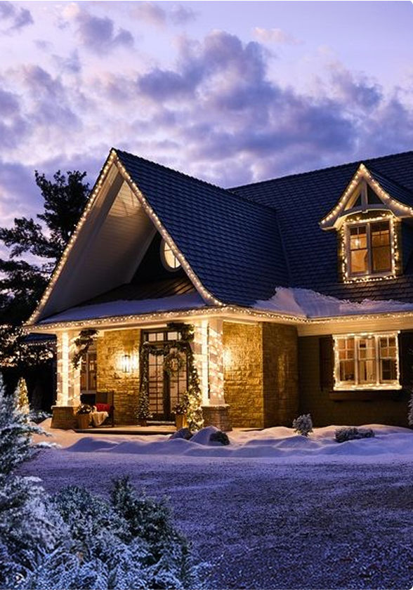 A two-storey home illuminated with warm white Christmas lights, creating a festive outdoor holiday display on a snowy evening.