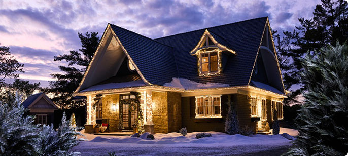A two-storey home illuminated with warm white Christmas lights, creating a festive outdoor holiday display on a snowy evening.