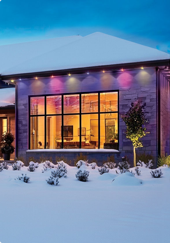 A modern home decorated with customizable multicolour smart Christmas lights along the roofline and exterior walls in a snowy setting.