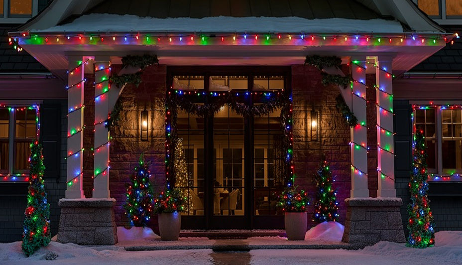 Home exterior decorated with multicolour Christmas lights along the roofline, columns and entryway in a snowy winter setting.