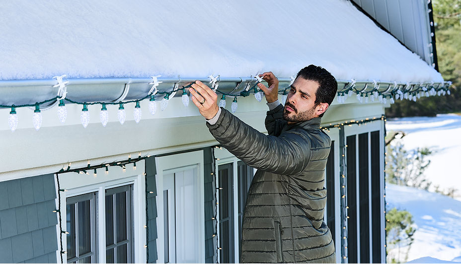 A person attaching white Christmas lights along the roofline of a snow-covered home.