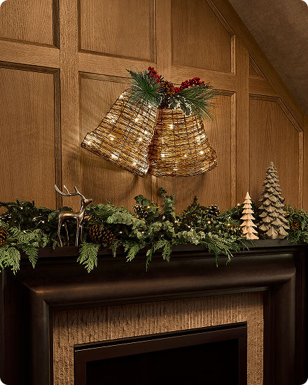 Pair of illuminated bell decorations displayed above a mantel with holiday greenery and pinecones.