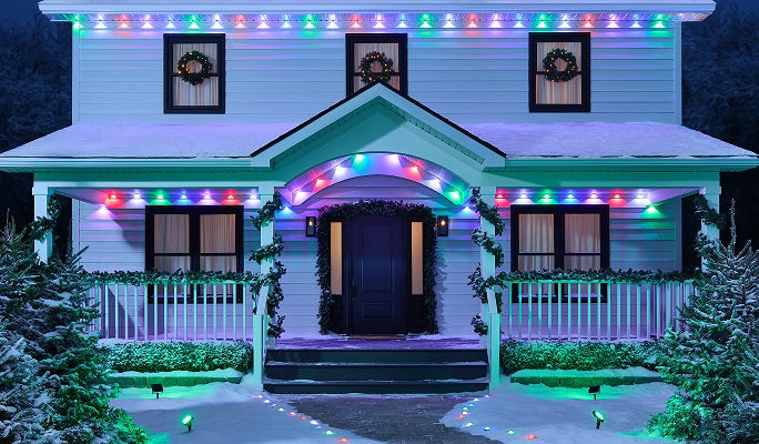 Home decorated with synchronized NOMA Smart Lights along the roofline, porch and pathway, creating a coordinated multi-colour holiday lighting display in a snowy outdoor setting.