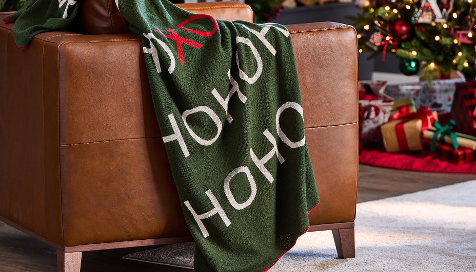 Une couverture verte avec les mots « HO HO XO » en lettres blanches, drapée sur un fauteuil en cuir brun, avec un sapin de Noël en arrière-plan.