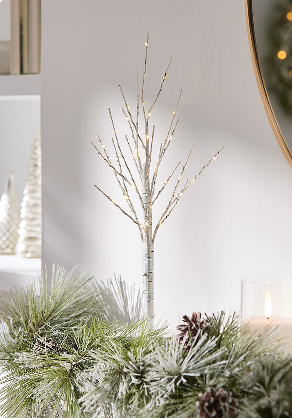 White birch-style tabletop tree featuring illuminated branches, displayed among frosted greenery and pinecones in a softly styled holiday vignette.