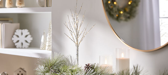 Pre-lit tabletop cedar tree displayed in a white ceramic pot on a side table, illuminated with warm white lights and styled alongside candles in a bright, neutral living space.