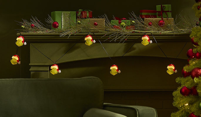 Grinch-themed HD string lights featuring illuminated Grinch faces draped across a fireplace mantel decorated with holiday greenery and wrapped gifts.