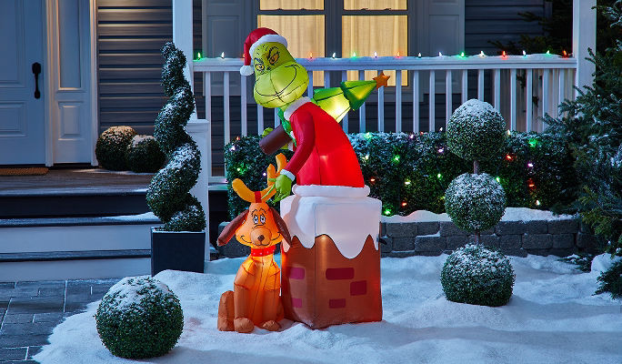 An Airblown Grinch climbing out if a chimney holding a Christmas tree with Max the dog beside it, displayed on a snowy front lawn.