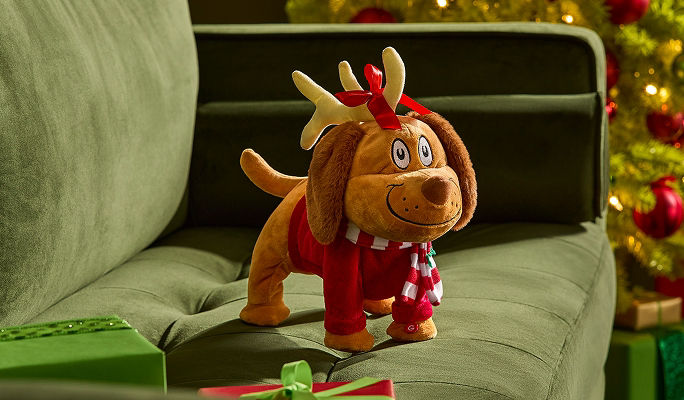 A Max the dog plush wearing a red sweater, striped scarf and reindeer antlers, displayed on a green sofa beside wrapped Christmas gifts.