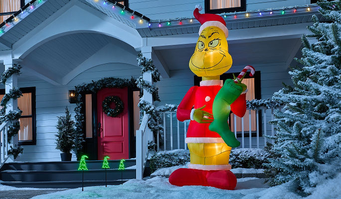 An Airblown Colossal Grinch inflatable holding a stocking filled with a candy cane, displayed on a snowy front lawn as part of an outdoor Christmas display.
